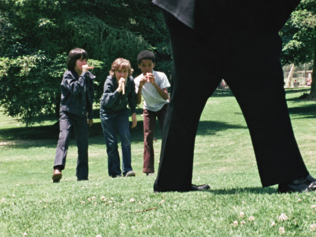 Restored archival video still of Ray Brown with two other boys playing instruments outdoors, with an adult’s trousered legs in the foreground.